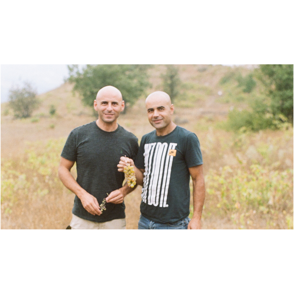 Winemakers Eddie Chami and Abdullah Richi standing in a field. Abdullah is holding a bunch of grapes. 